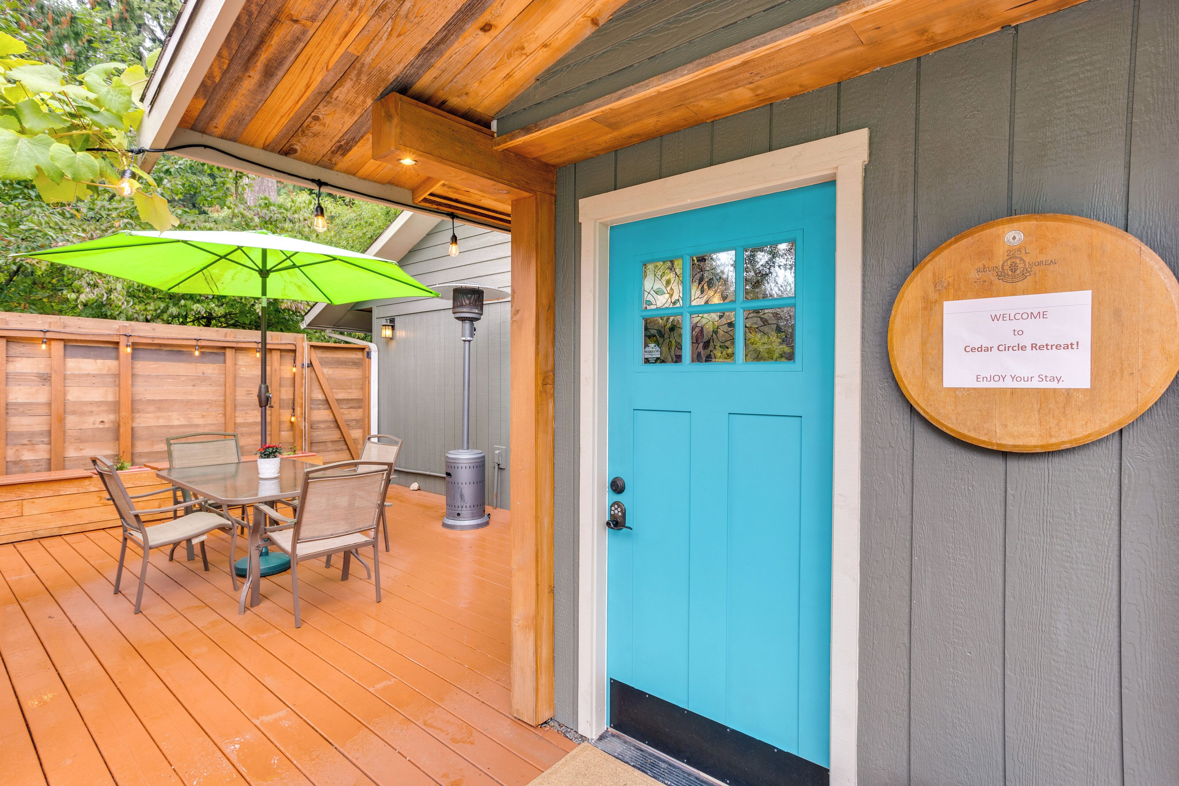 A wooden deck with a table and chairs, a green umbrella, and a blue door with a welcome sign.
