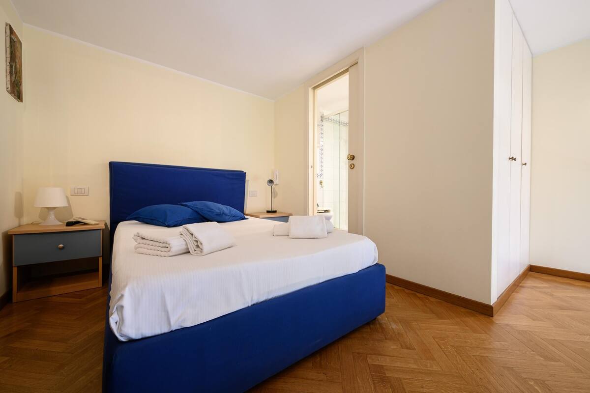 Une chambre à coucher avec un grand lit, une table de chevet, une lampe et une porte donnant sur une autre pièce.