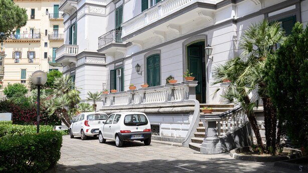 Ein weißes Gebäude mit grünen Fensterläden und einem Balkon, geparkte Autos und Palmen.