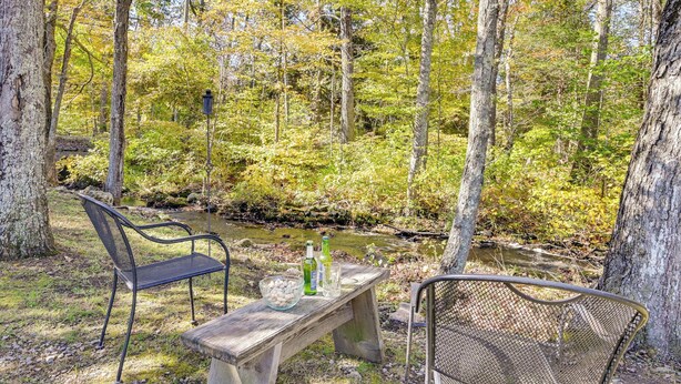 A wooden bench with a metal chair, a small table with bottles and glasses, and a view of a forested area with a stream. - Quiet Killingworth Rental w/ Deck & Grill! (Killingworth)