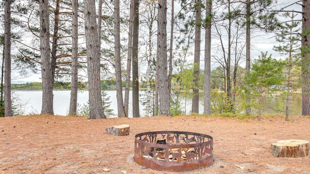 A rustic campsite with a metal fire pit surrounded by pine trees and a lake in the background. - Boat Dock & Fire Pit: Lakefront Monico Home! (Rhinelander)