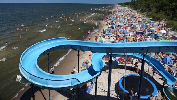 A water park with a large blue water slide and a sandy beach area with umbrellas and people.
