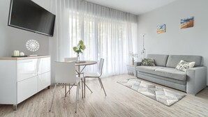 A modern living room with a grey sofa, a dining table with chairs, a TV mounted on the wall, and a large window with sheer curtains.