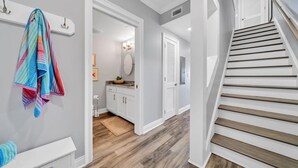 A hallway with a wooden floor, a staircase with wooden steps, and a bathroom with white cabinets and a towel hanging on the wall.