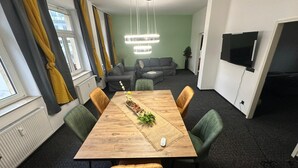 Moderni olohuone, jossa on puinen ruokapöytä, vihreät tuolit, harmaa sohva ja seinälle asennettu televisio.