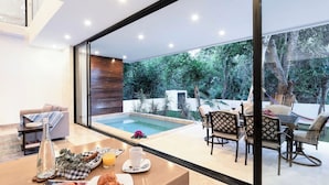 A modern outdoor dining area with a pool, comfortable seating, and a view of lush greenery.