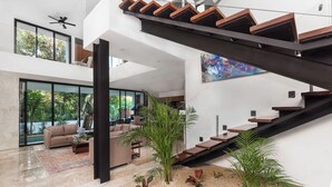 A spacious living area with a modern staircase, large windows, and a view of greenery outside.