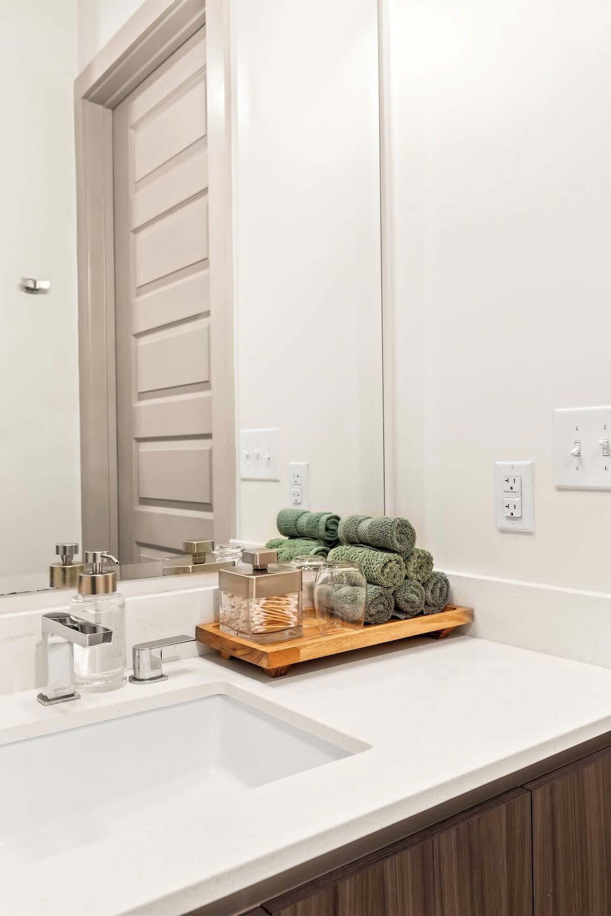 A modern bathroom with a white sink, a mirror, and a wooden tray holding rolled towels and toiletries.