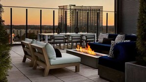 Eine Dachterrasse mit Sitzgelegenheiten im Freien, einer Feuerstelle und Blick auf die Stadt im Sonnenuntergang.