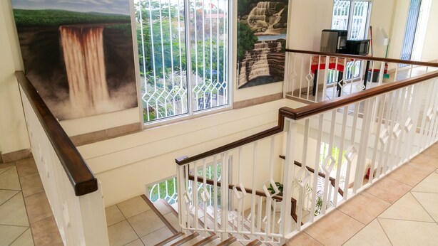 A staircase with white railings and a wooden handrail, leading to a balcony with a view of greenery and a waterfall painting. - King Studio - One
 (Georgetown)