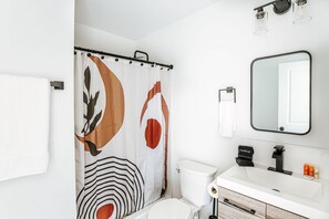 Une salle de bain moderne avec un rideau de douche orné de motifs abstraits, des toilettes, un lavabo avec un miroir et un porte-serviettes.