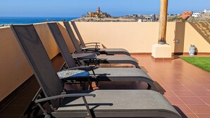 A row of sun loungers on a terrace with a view of the sea and buildings in the background.