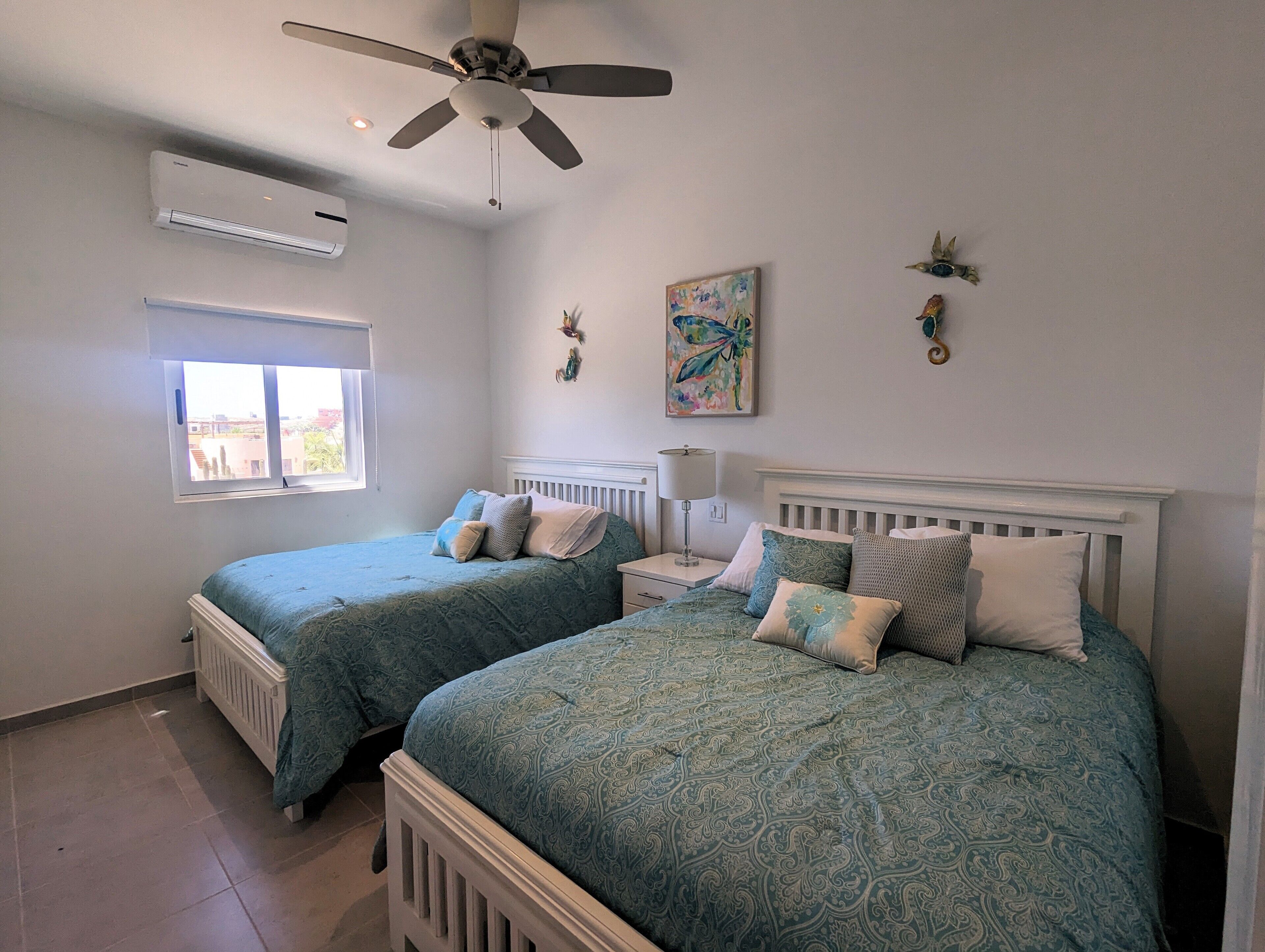 A bedroom with two twin beds, a ceiling fan, an air conditioning unit, a window, and a wall-mounted lamp.