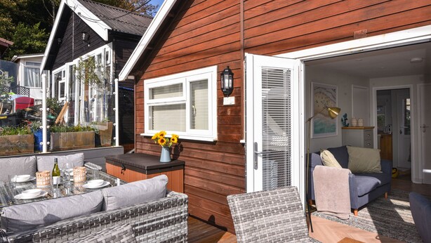 Une maison en bois dotée d’un patio aménagé avec des meubles en osier, une table à manger et un vase rempli de fleurs.