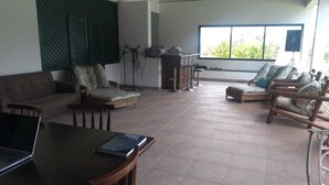 A spacious living area with a sofa, armchairs, a dining table with a laptop, and a large window with a view of greenery.