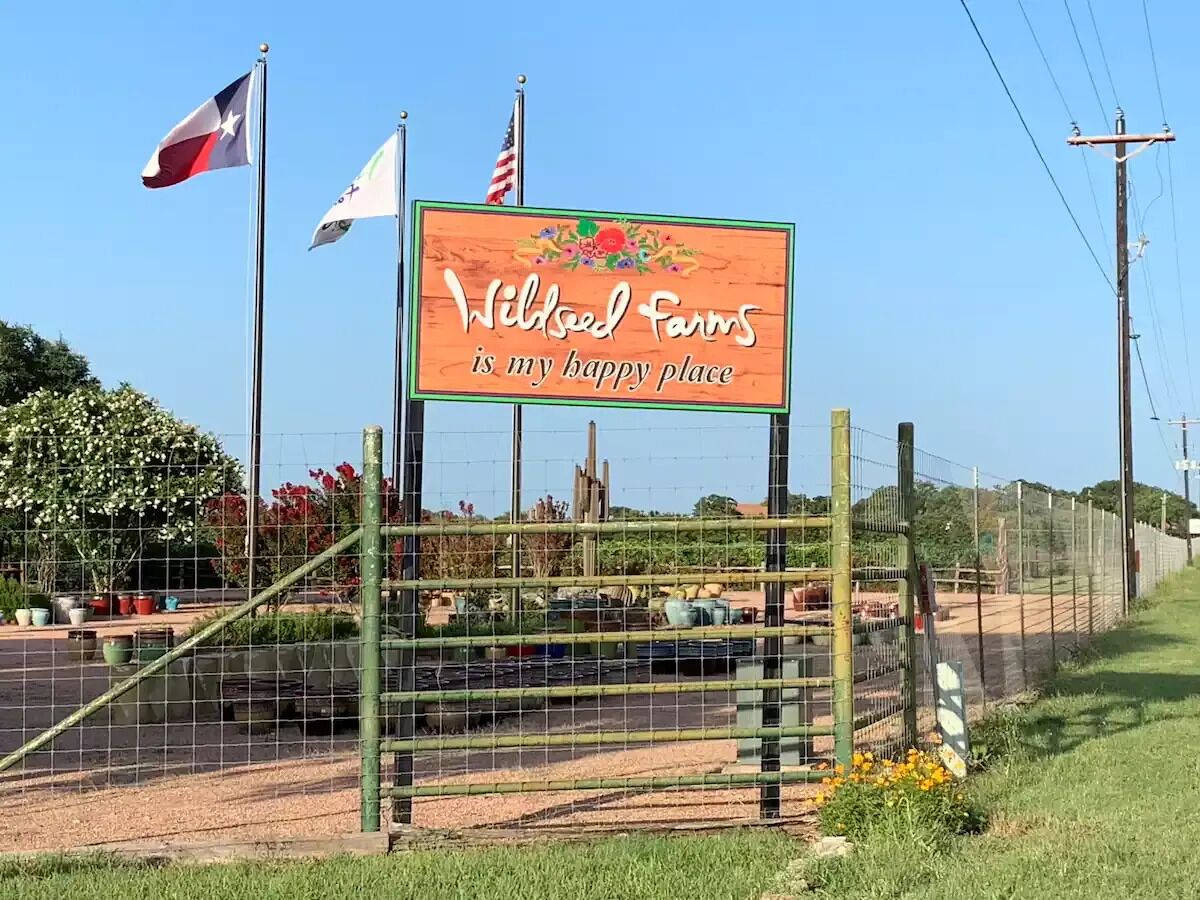 Une pancarte sur laquelle on peut lire : « Wildseed Farms, mon havre de paix », avec une clôture et des drapeaux en arrière-plan.