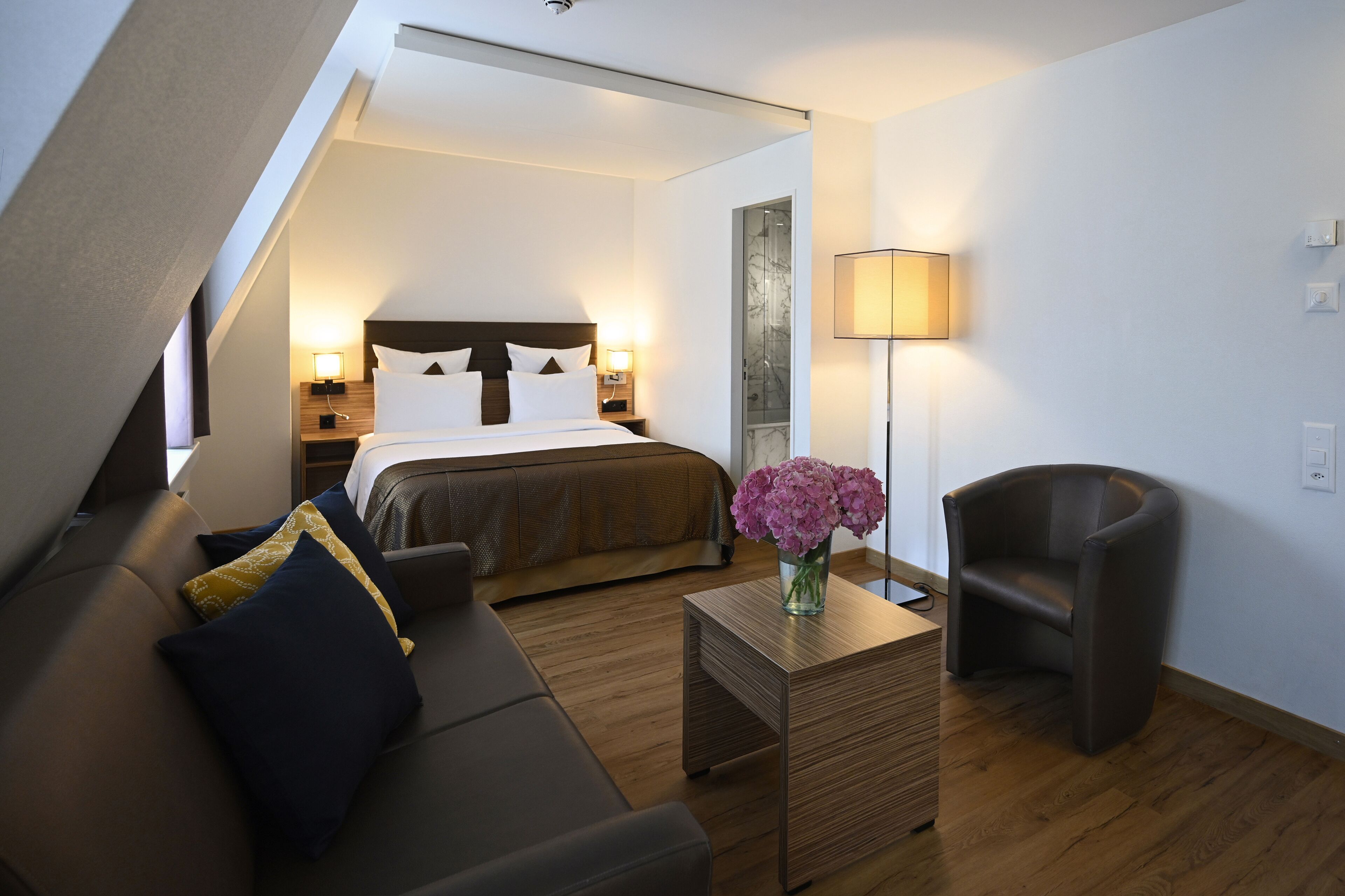 A modern hotel room with a bed, a sofa, a chair, a small table with a vase of flowers, and a lamp.