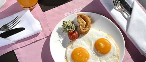 Un plato de desayuno con dos huevos fritos, un tomate pequeño y un pastel sobre un plato blanco, acompañado por un tenedor y un cuchillo sobre una servilleta rosa, con un vaso de jugo de naranja al fondo.