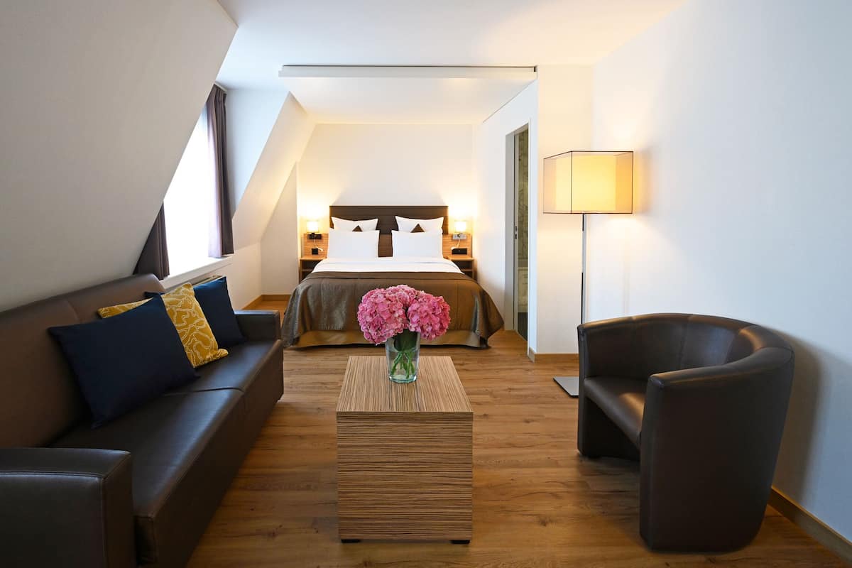 A modern hotel room with a loft-style ceiling, a large bed, a sofa, a chair, and a bedside table with a vase of flowers.
