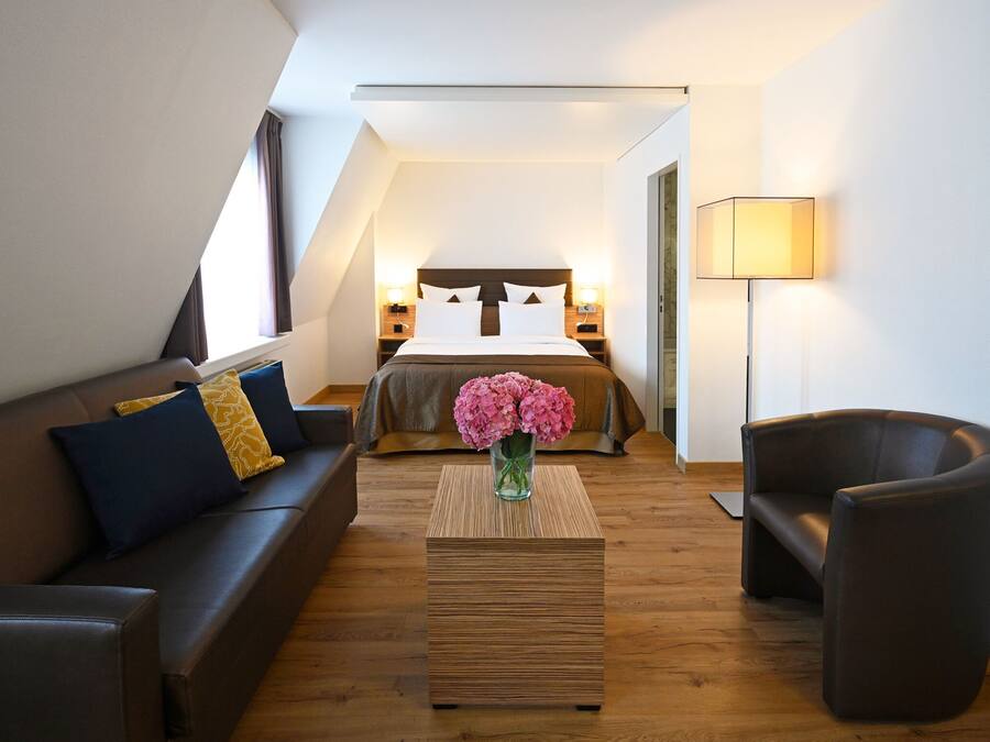 Ett modernt hotellrum med ett tak i loftstil, en stor säng, en soffa, en fåtölj och ett sängbord med en vas med blommor.