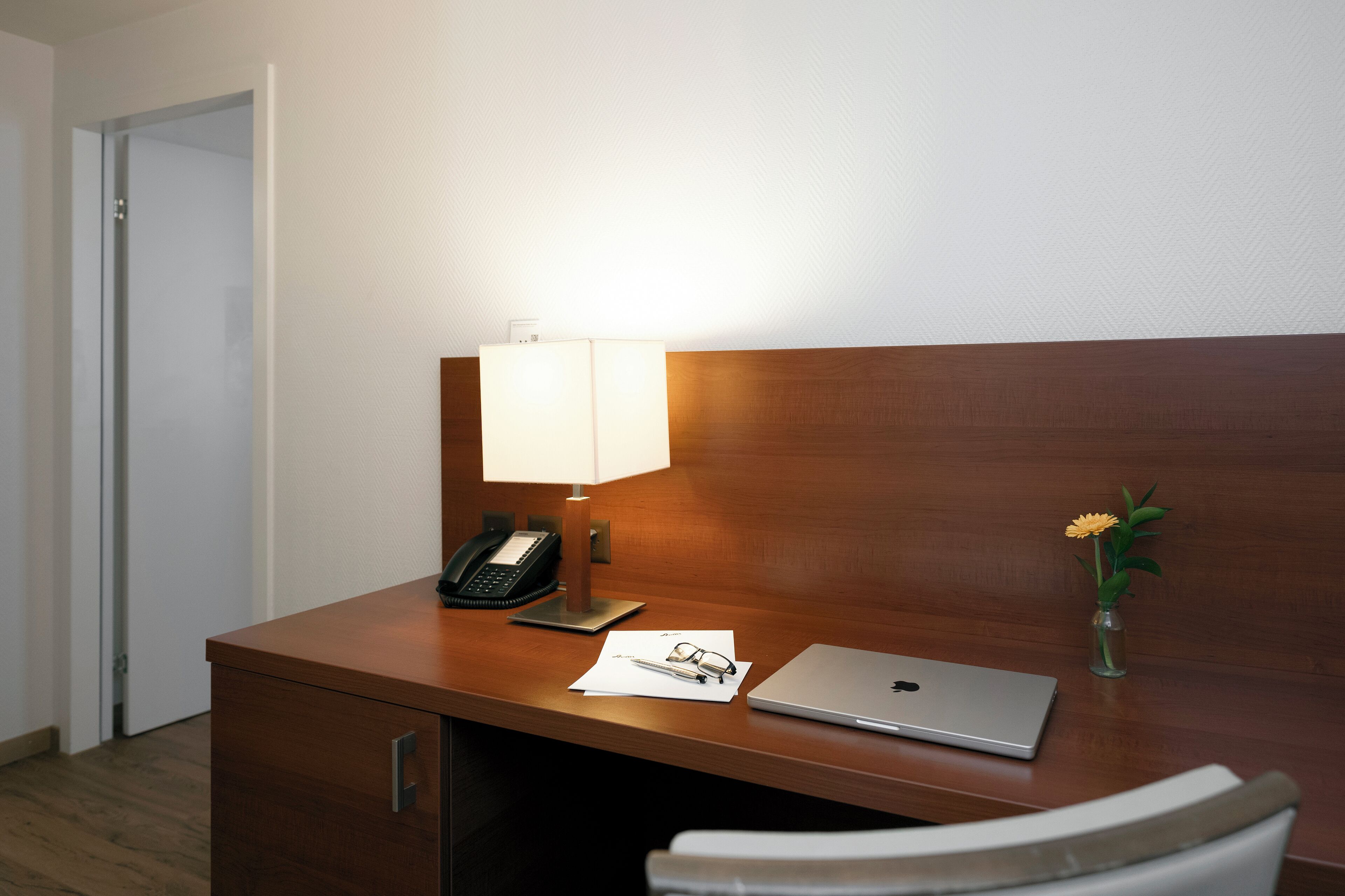Escritorio de habitación de hotel con una computadora portátil, una lámpara, un teléfono, un florero con flores y una silla.