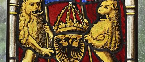 A stained glass window with two lions holding a shield and a crown, set within an arched frame.