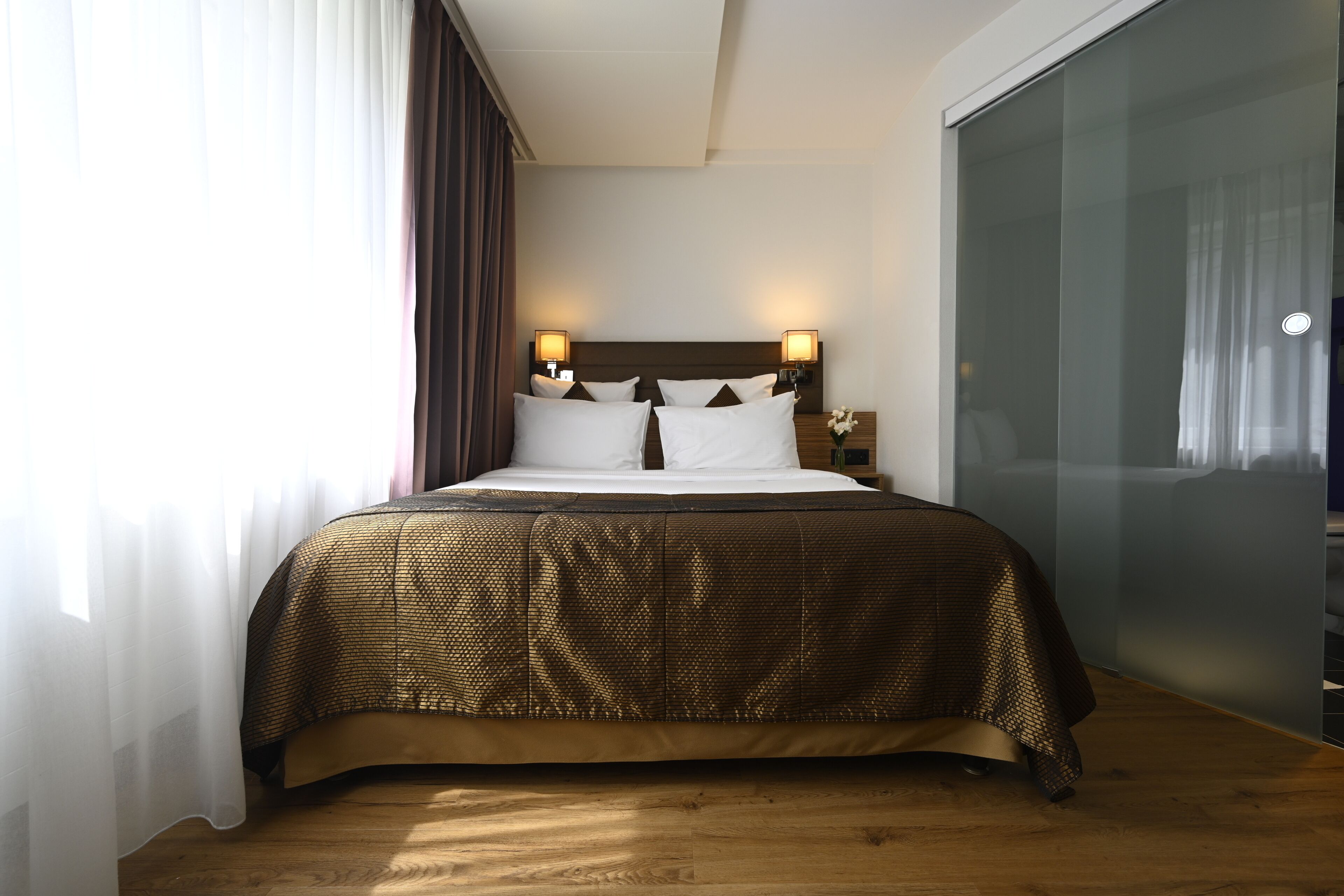 A hotel room with a large bed, two bedside lamps, a wooden headboard, and a view of the city through a glass door.