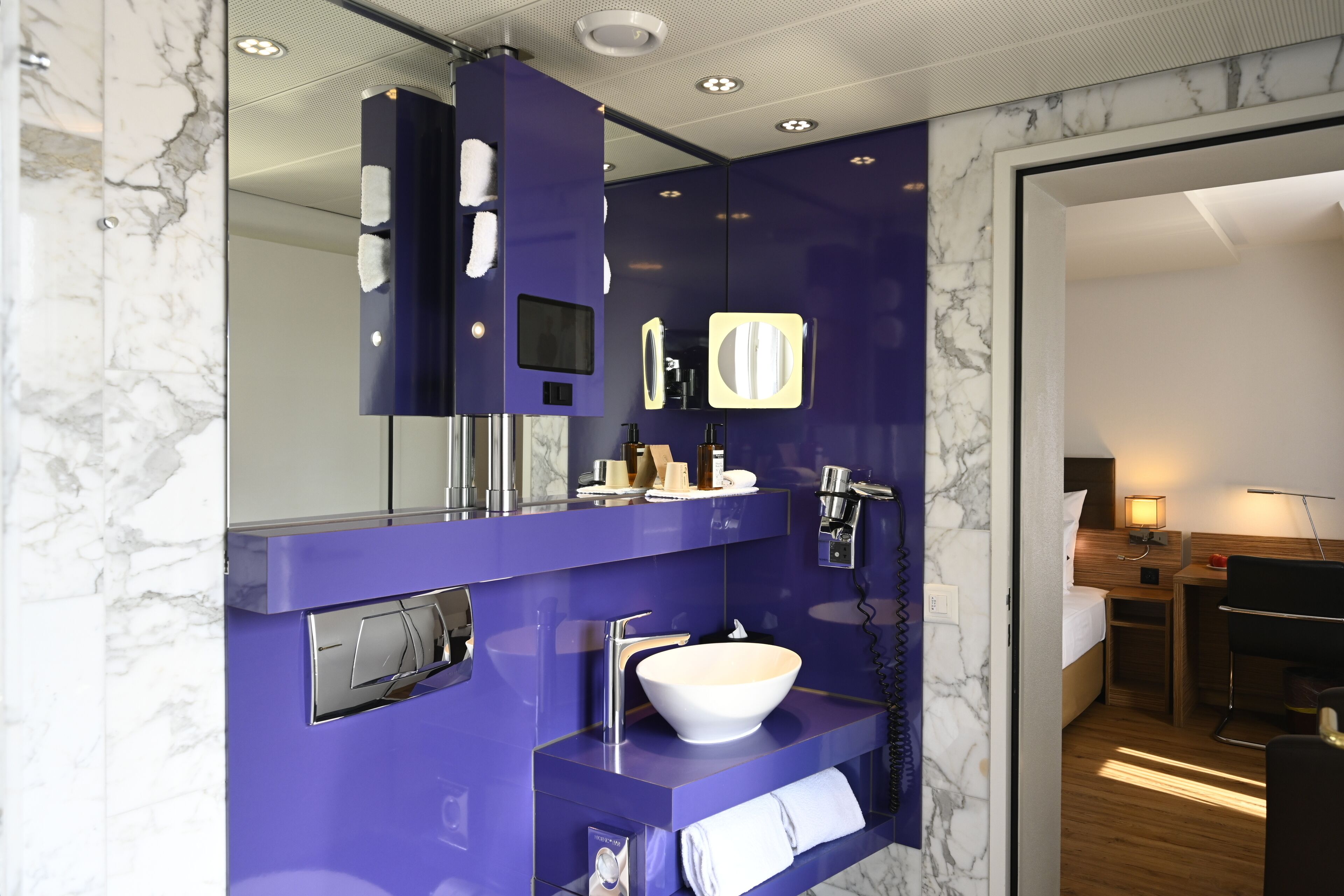 Une salle de bain moderne avec un meuble-lavabo violet, un lavabo blanc et un miroir. Une petite table avec une lampe et une chaise se trouvent dans la pièce adjacente.