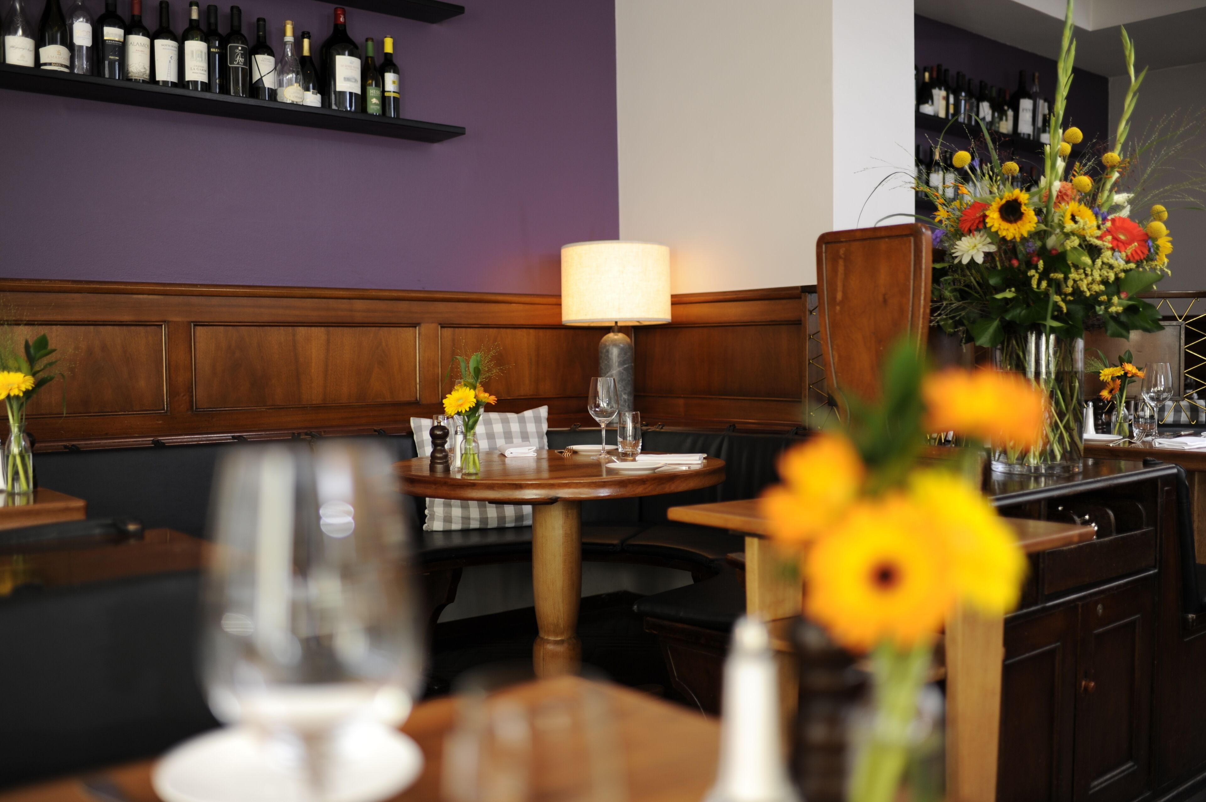Interior de restaurante com mesas de madeira, parede roxa e vários vasos com flores.