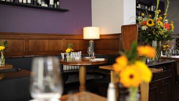 Interior de restaurante com mesas de madeira, parede roxa e vários vasos com flores.