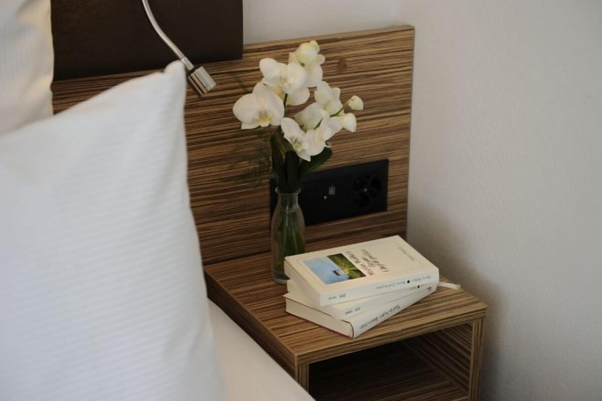Una mesita de noche de madera con un jarrón blanco lleno de flores blancas, un libro y un enchufe.