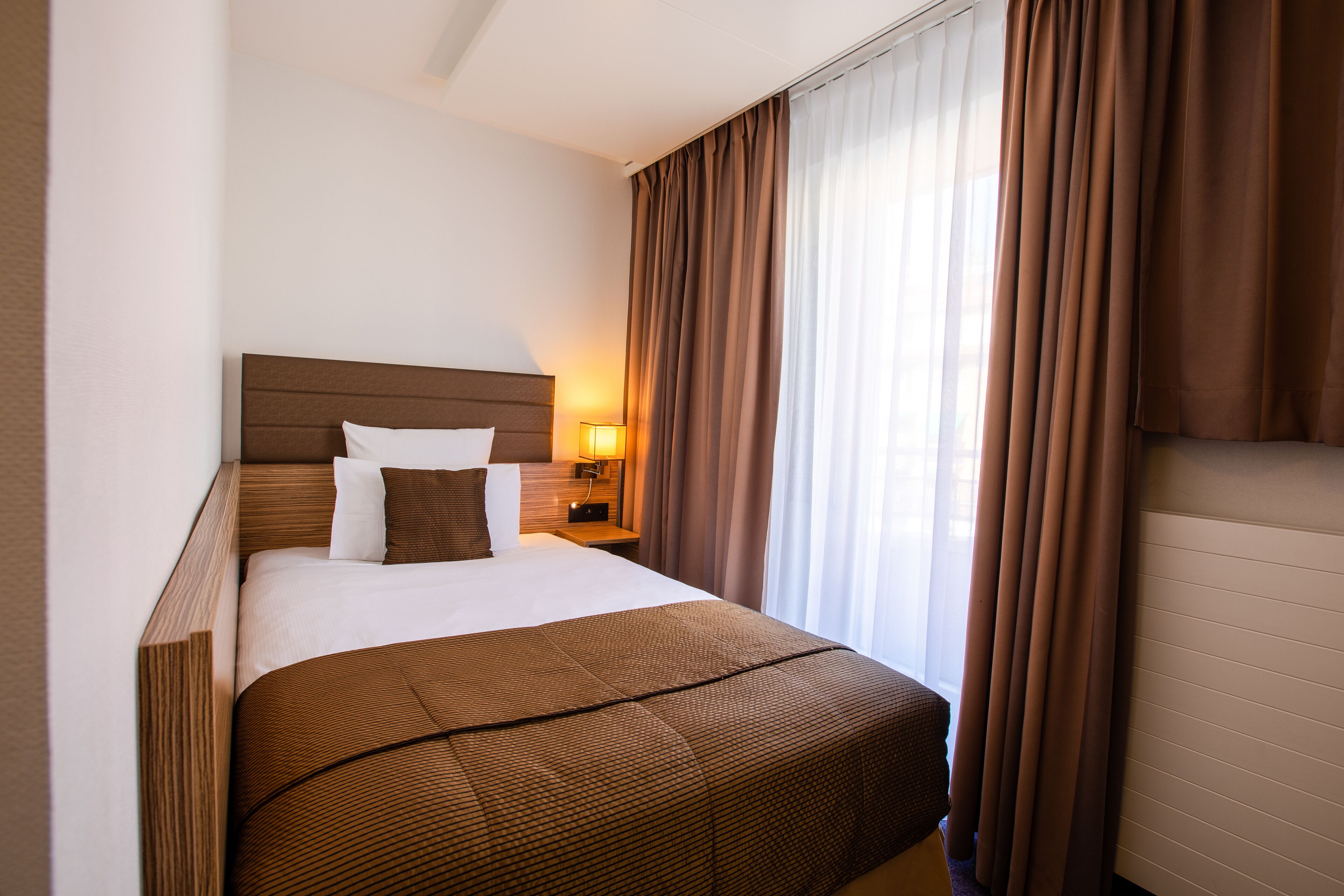 Una habitación de hotel con una cama grande, una mesita de noche con lámpara y una ventana con cortinas.