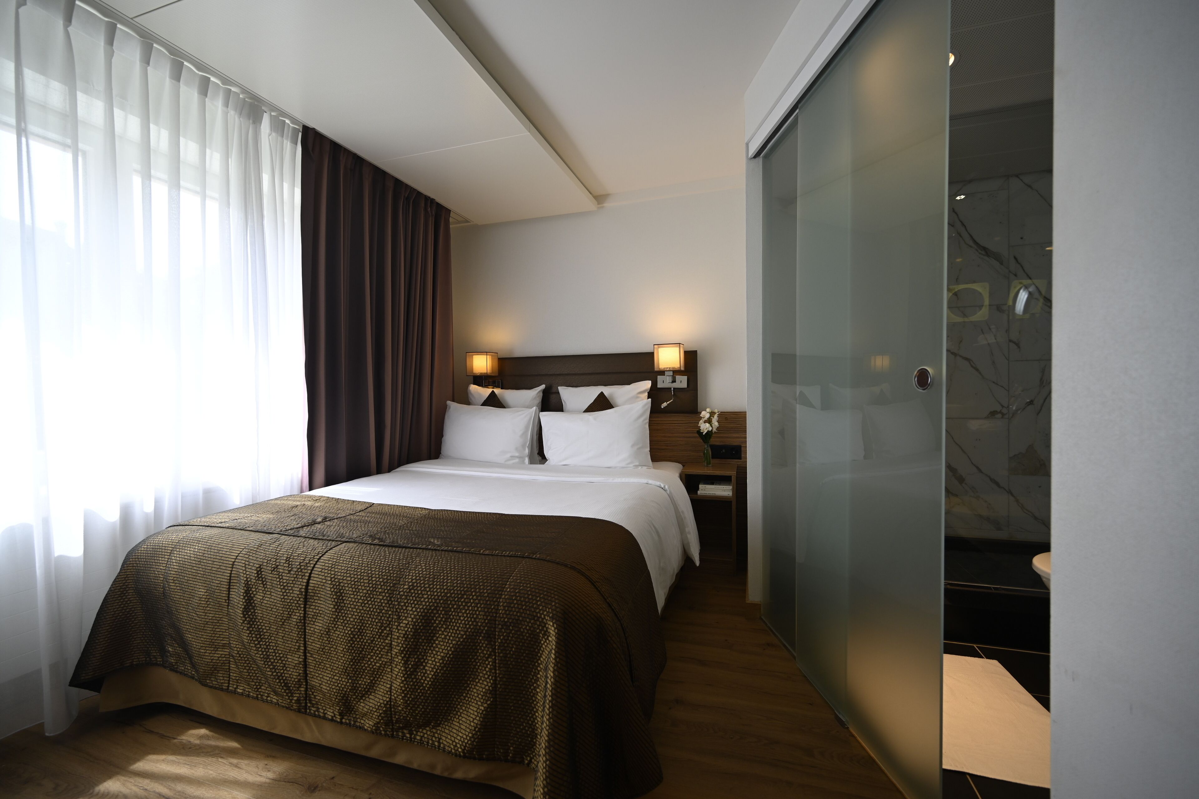 Une chambre d’hôtel avec un grand lit, une table de chevet, une cabine de douche en verre et une vue sur l’extérieur à travers des rideaux transparents.