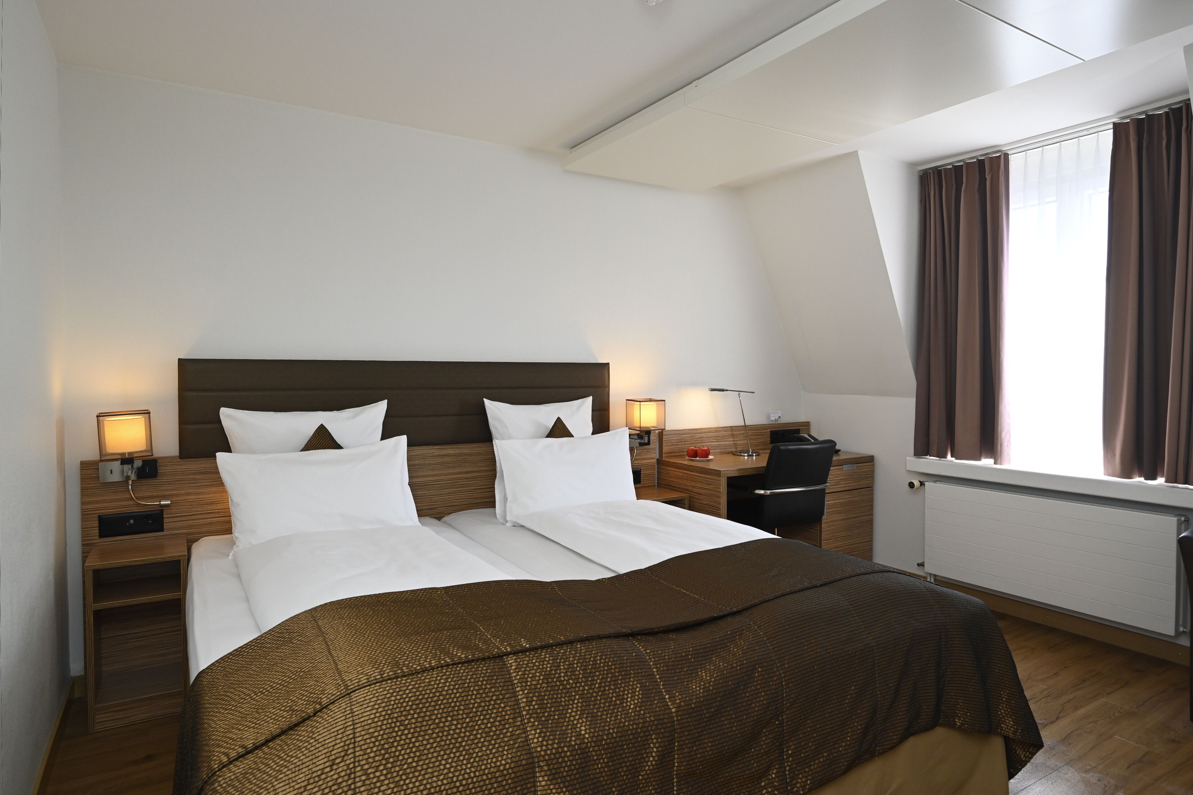 A hotel room with a large bed, two bedside tables, a desk, and a window with curtains.