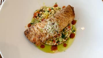 Un plato con un trozo de pescado a la parrilla, una ensalada de cereales y una salsa verde.