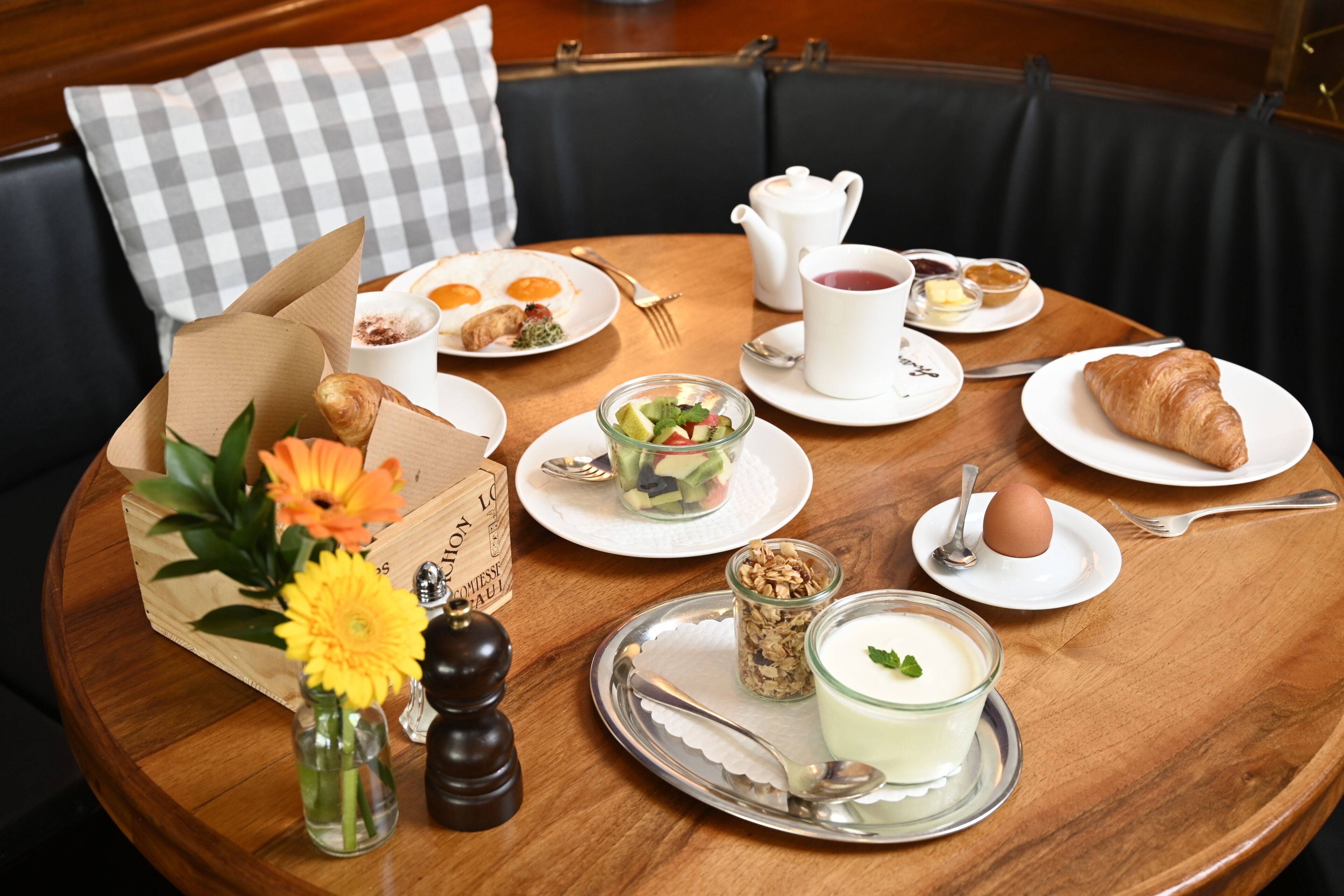 Een ronde eettafel met diverse ontbijtartikelen, zoals croissants, eieren en een fruitsalade. Ook aanwezig zijn sauzen, een theepot en een vaas met bloemen.