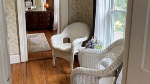 A room with a wooden floor, white wicker chairs, a small rug, and a view of the outdoors through a window with sheer curtains.
