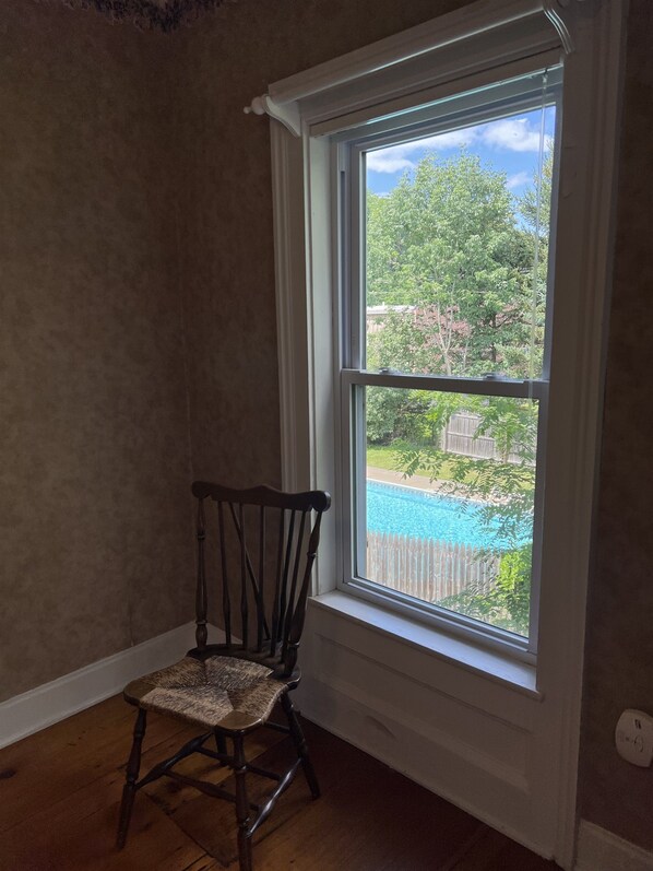 Una habitación con una silla de madera, una ventana con vistas a una piscina y una pared blanca.