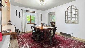 Un coin repas avec une table en bois, des chaises noires, un tapis à motifs rouges, une fenêtre avec des rideaux et un luminaire fixé au mur.