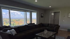 A modern living room with a sectional sofa, a coffee table, and a view of autumn foliage through large windows. - In ground Pool with unbeatable views, ASV (Otego)