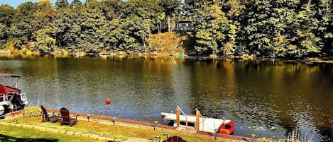 A lakeside view with a dock, chairs, and a house in the background.