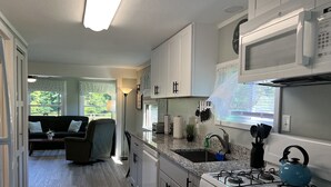 A modern kitchen with white cabinets, a gas stove, a microwave, and a sink. There is a sofa and a floor lamp in the adjacent living area.