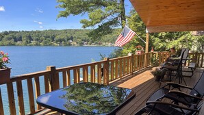 A wooden deck with outdoor furniture overlooking a lake, an American flag, and a view of a forested area. - Lonestar - New build, stunning lake views, near CDP (Milford)