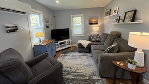 Ein modernes Wohnzimmer mit einem grauen Sofa, einem Fernseher auf einem weißen Schrank, einem kleinen Tisch mit einer Lampe und einem dekorativen Regal mit Bildern.