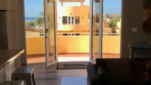 Una sala moderna con un sofá, una mesa de centro y una zona de cocina con taburetes. Hay un amplio ventanal corredero que da a un balcón con vistas al océano y a edificios.