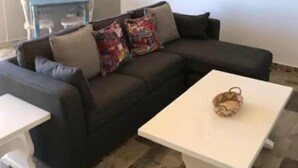 Un salón con un sofá gris oscuro, cojines coloridos, una mesa de centro blanca y dos cuadros en las paredes.