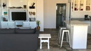 Una sala de estar moderna con un sofá gris, una mesita blanca y un televisor sobre un mueble blanco. Se ve un ventilador de techo y el piso es de baldosas.