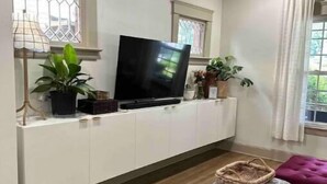 A living room with a TV on a white cabinet, wooden flooring, a purple sofa, and a window with curtains. - Cleveland Hideaway on Clifton (Cleveland)