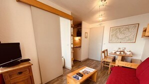 Ett kompakt vardagsrum med en tv på ett träskåp, ett träbord med tidningar, en röd soffa, ett trämatbord med stolar och ett köksområde i bakgrunden.