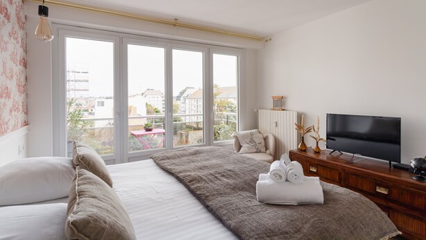 A bedroom with a large bed, a TV, and a view of the city through large windows. - Starfish - Charmant appt à 3min à pied de la plage (Saint-Malo)